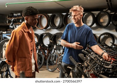 Young man explaining bicycle features to Black young man in bike shop, both standing among rows of bicycles with wheels displayed on wall in background