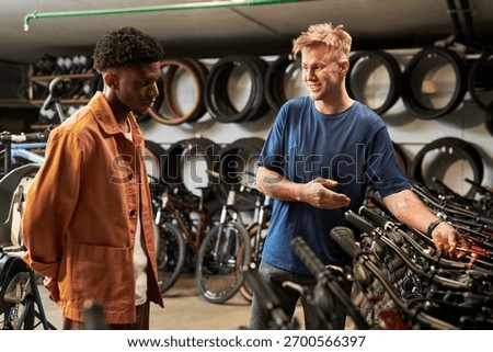 Young man explaining bicycle features to Black young man in bike shop, both standing among rows of bicycles with wheels displayed on wall in background