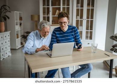 Young man explaining digital technology to an older man, both looking intently at a laptop screen, fostering inter generational connection and learning in a bright home environment