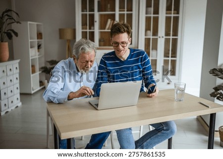 Young man explaining digital technology to an older man, both looking intently at a laptop screen, fostering inter generational connection and learning in a bright home environment