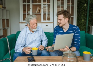 Young man explaining how to use a laptop to an attentive senior man while sitting on a green sofa, establishing an inter generational connection and sharing knowledge