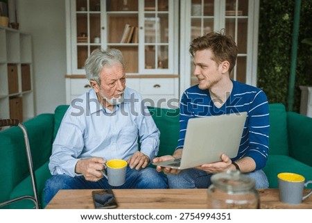 Young man explaining how to use a laptop to an attentive senior man while sitting on a green sofa, establishing an inter generational connection and sharing knowledge