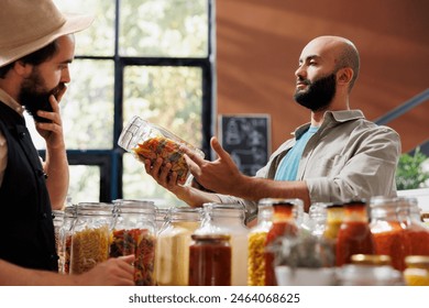 A young man explores an eco-friendly store, looking at a variety of fresh organic pantry products in reusable packaging. A Middle Eastern client examines jars of pasta and grains. - Powered by Shutterstock