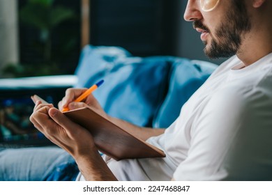 Young man with eye patches studying from home writing in a notebook with a pencil, sitting on a sofa. Sitting in living room in the evening. Student studying - Powered by Shutterstock