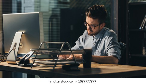Young man in eyeglasses doing overwork project sitting alone in office with lamplight.  - Shutterstock ID 1099777979