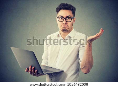Young man in eyeglasses holding laptop and looking at camera in misunderstand shrugging with shoulders. 