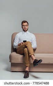 young man in eyeglasses holding smartphone and looking at camera while sitting on sofa isolated on grey