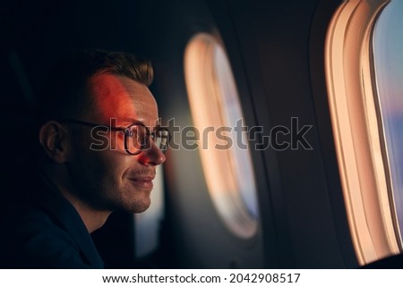 Young man with eyeglasses traveling by airplane. Passenger looking through window during flight at sunset.

