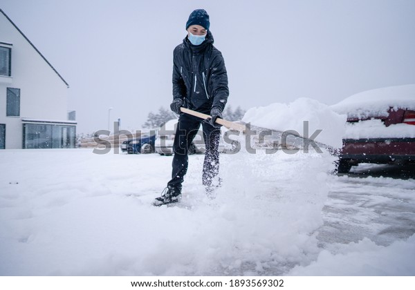 Young man with face\
mask holding shovel and shoveling fresh white snow at yard of house\
during blizzard in winter day. Man on parking with shovel helps\
cars stuck in snow\
