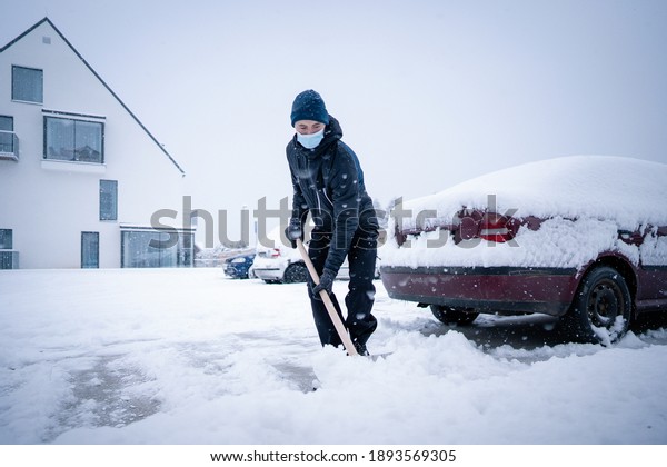 Young man with face\
mask holding shovel and shoveling fresh white snow at yard of house\
during blizzard in winter day. Man on parking with shovel helps\
cars stuck in snow\
