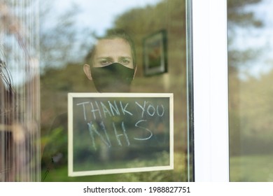 A Young Man With A Face Mask On Looking Out The Window During His Self-isolation At Home, He Is Holding Up A Sign With Thank You NHS Written On It