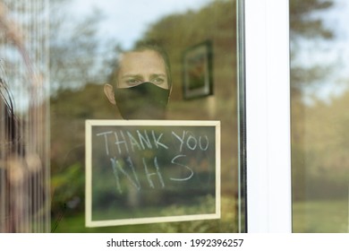 A Young Man With A Face Mask On Looking Out The Window During His Self-isolation At Home, He Is Holding Up A Sign With Thank You NHS Written On It