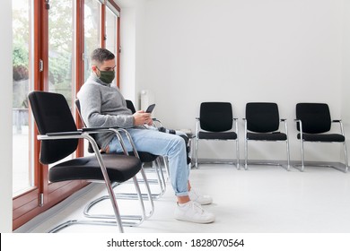 Young man with face mask sitting in a waiting room of a hospital or office looking at smartphone - focus on the man - Powered by Shutterstock