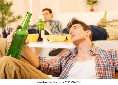 A Young Man With A Face Out Of Focus, Tired Of The Party Sitting On The Floor Leaning Against The Table, Holding A Bottle Of Beer. In The Background You Can See Two Hungover Guy Lying On The Couch.