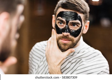 Young man with facial mask looking in mirror