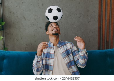 Young man fan in casual clothes shirt cheer up support football team sit on blue sofa hold soccer ball on head soccer ball do winner gesture rest watch tv indoors room grey wall. Sport leisure concept - Powered by Shutterstock