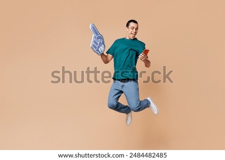 Young man fan wear basic blue t-shirt use mobile cell phone cheer up support football sport team hold soccer ball, foam 1 fan glove finger up, watch tv live stream isolated on plain beige background