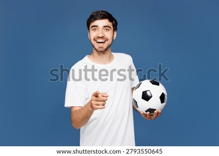 Young man fan wear basic white t-shirt cheer up support football sport team hold in hand soccer ball watch tv live stream point index finger camera on you motivating isolated on plain blue background