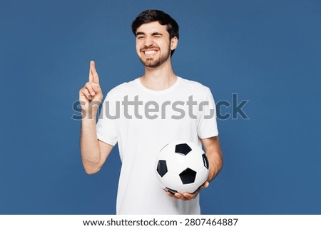 Young man fan wear basic white t-shirt cheer up support football sport team hold in hand soccer ball watch tv live stream wait special moment, keeping fingers crossed isolated on plain blue background