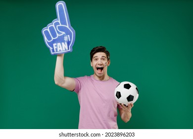 Young man fan wears pink t-shirt foam 1 one sign glove finger cheer up support football sport team hold in hand soccer ball watch tv live stream scream isolated on dark green color background studio