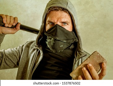 Young Man As Fanatic And Aggressive Ultra Anarchist Rioter . Furious Anti-system Protester In Face Mask Holding Baton Throwing Brick Looking Hostile Fighting At Violent Riot In Radical Demonstration