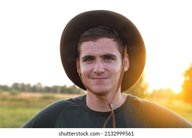 Young man farmer in cowboy hat at agricultural field on sunset with sun flare. Closeup portrait of millennial man with hat, standing on nature background, outdoors. Rancher. 