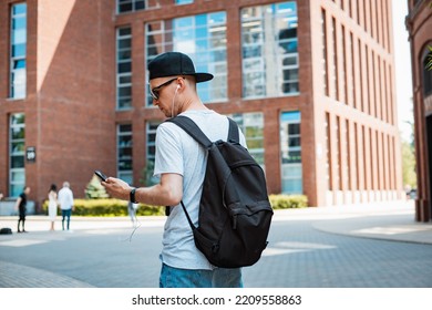 young man fashionable stylish guy sunglasses black cap walking around city holding a smartphone his hand and listening music headphones. Tourism, summer vacation, rear view, back view, backpack - Powered by Shutterstock