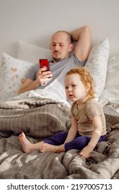 Young Man Father Laying On The Bed Using Cellphone With His Toddler Child Sitting On Foreground And Looking Disappointed. Kid Feeling Lonely. Selective Focus.