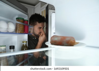 Young Man Feeling Bad Smell Of Spoiled Sausage In Refrigerator, View From Inside