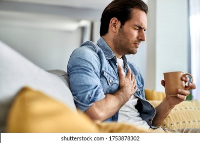 Young man feeling sick and holding his chest in pain while drinking tea in the living room.  - Shutterstock ID 1753878302