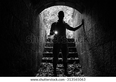 Young man with a flashlight enters the stone tunnel and looks in the dark, monochrome photo