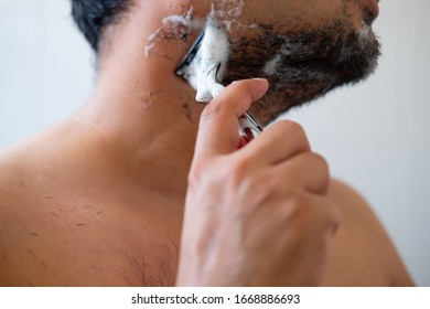 Young man with foam soap on his cheeks is standing and removing mustache by shaving from his face. - Shutterstock ID 1668886693