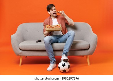 Young man football fan in shirt support favorite team with soccer ball sit on sofa at home watch tv live stream eat biting pizza isolated on orange background. People sport leisure lifestyle concept.