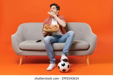 Young man football fan in shirt support favorite team with soccer ball sit on sofa at home watch tv live stream eat pizza dinner isolated on orange background. People sport leisure lifestyle concept.