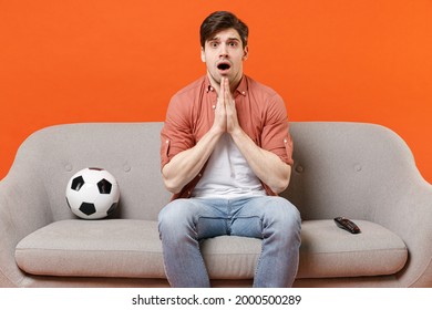 Young Man Football Fan In Shirt Support Team With Soccer Ball Sit Sofa Home Watch Tv Live Stream Hands Folded In Prayer Gesture, Begging Isolated On Orange Background Portrait People Sport Concept.