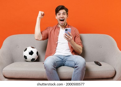 Young Man Football Fan In Shirt Support Team With Soccer Ball Sitting On Sofa Home Watching Tv Live Stream Use Mobile Cell Phone Do Winner Gesture Isolated On Orange Background. People Sport Concept.