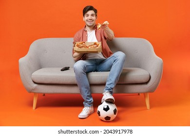 Young man football fan in shirt support favorite team with soccer ball sit on sofa at home watch tv live stream give slice of pizza isolated on orange background People sport leisure lifestyle concept