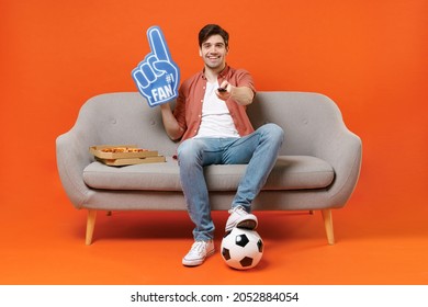 Young man football fan in shirt support team with soccer ball sit on home sofa watching tv live stream hold pizza foam glove finger switch channel isolated on orange background People sport concept - Shutterstock ID 2052884054