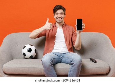 Young man football fan in shirt support team with soccer ball sit sofa home watch tv use mobile cell phone blank screen workspace area show thumb up isolated on orange background People sport concept.