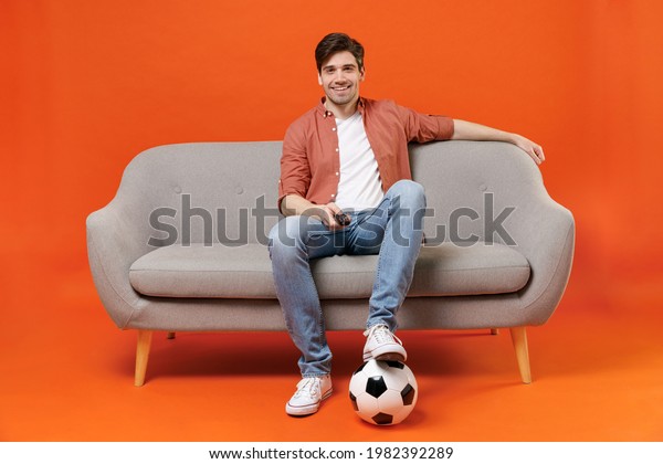 Young man football fan in shirt support favorite\
team with soccer ball sit on sofa at home watch tv live stream\
switch channel isolated on orange background. People sport leisure\
lifestyle concept.