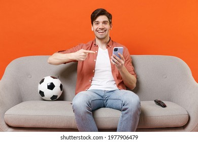 Young Man Football Fan Wearing Shirt Support Team With Soccer Ball Sitting On Sofa Home Watching Tv Live Point Index Finger On Mobile Cell Phone Isolated On Orange Background. People Sport Concept.