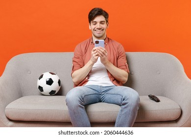 Young Man Football Fan Wearing Shirt Support Team With Soccer Ball Sitting On Sofa Home Watching Tv Live Using Mobile Cell Phone Makes Bets Isolated On Orange Background Studio. People Sport Concept.