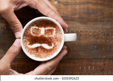 young man forming a heart with his hands around a cup of cappuccino, with a pair of eyeglasses and a moustache drawn with cocoa powder on its foam, on a wooden table with some blank space on the right