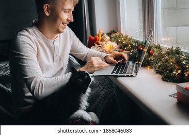 Young man freelancer in pajama working distance online having video call on laptop computer with cat at home office. Christmas mood, New Year decoration, xmas vacation. Lockdown isolation.