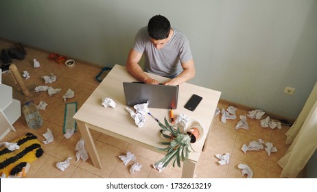 A young man, the freelancer sits at a Desk and working with laptop at home. Around him, the chaos and strong mess. Dirty unkempt apartment, all in the garbage