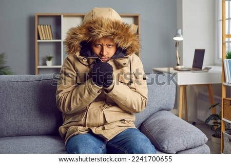 Young man freezing indoors without central heating. African American man wearing warm winter clothes sitting on sofa in very cold room at home looking at camera with angry disappointed face expression