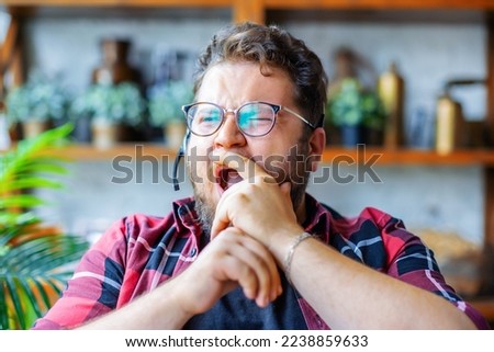 A young man in front of a webcam during an online video call. He yawns and covers his mouth with his hand. The concept of burnout from the same type of work and overworking.