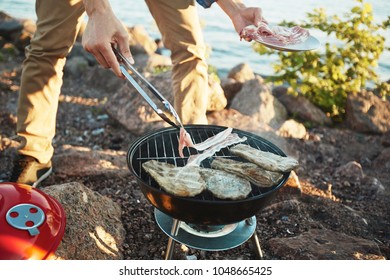 Young Man Frying Bacon And Meat On Grill During Picnic By Seaside