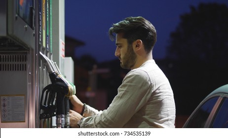 Young Man Fueling His Car At Gas Station, Using The Pump Nozzle. Late At Night