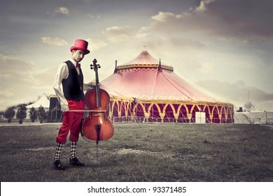 Young Man In Funny Clothes Holding A Cello With Circus In The Background